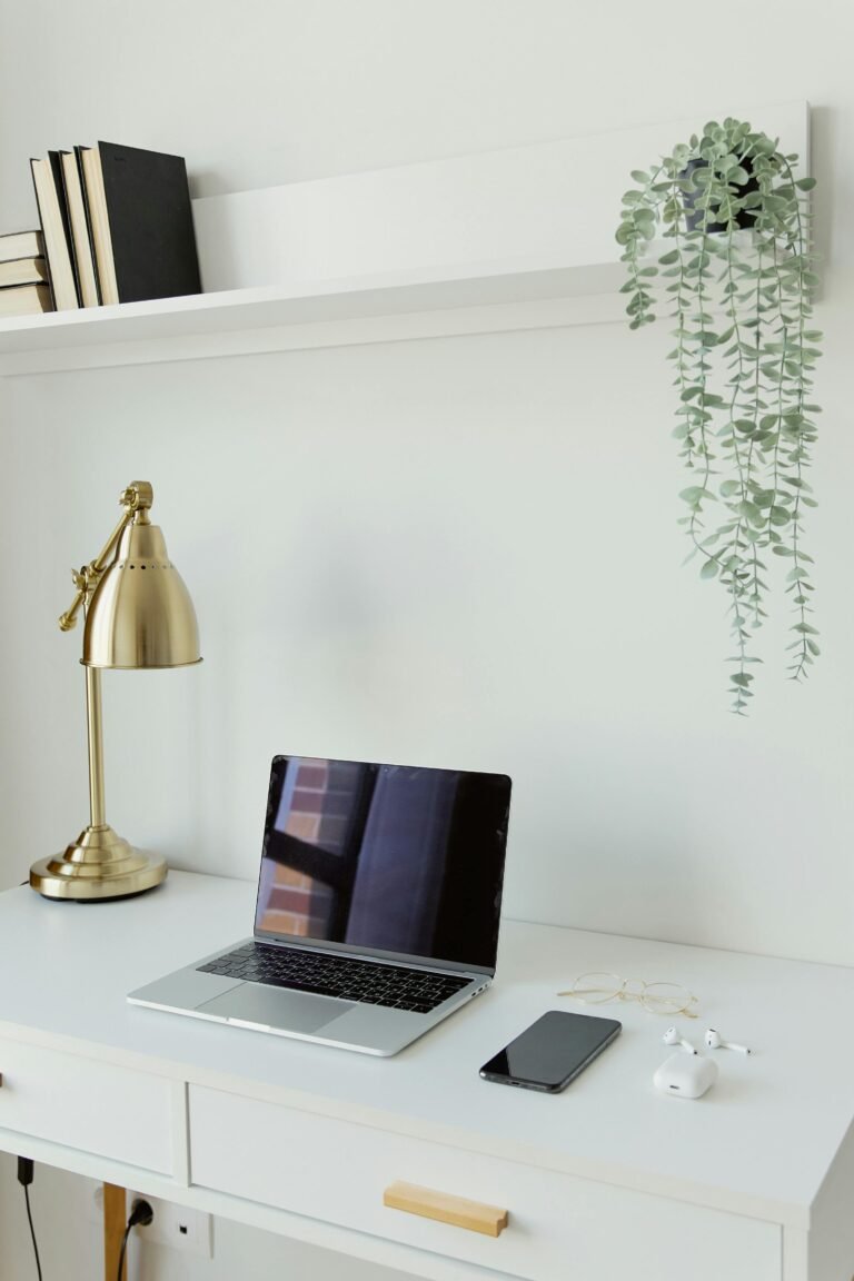 A clear, minimal white small desk with a laptop and a wall shelf above - how to free up space on a small desk by keeping essentials minimal and going vertical.