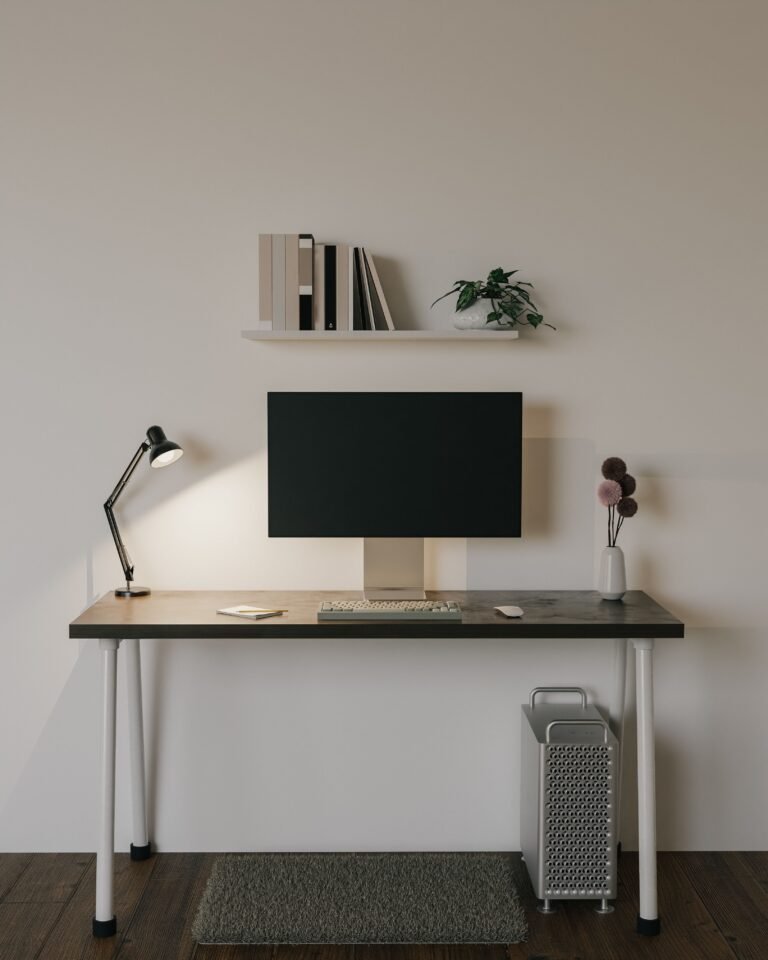 A minimalist slab desk with no drawers against a white wall — a shelf above holds books upright and a small plant, with a monitor, keyboard and lamp on a clear, tidy surface below — showing how to organize a small desk without drawers using vertical space.