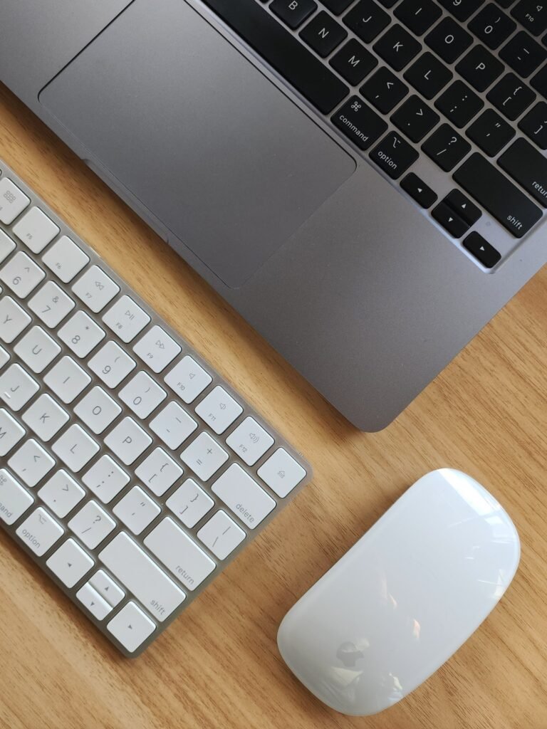 A compact wireless keyboard and a white mouse side by side on a wooden desk next to a laptop