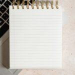Blank spiral notepad resting on a laptop keyboard, seen from above on a speckled cream desk