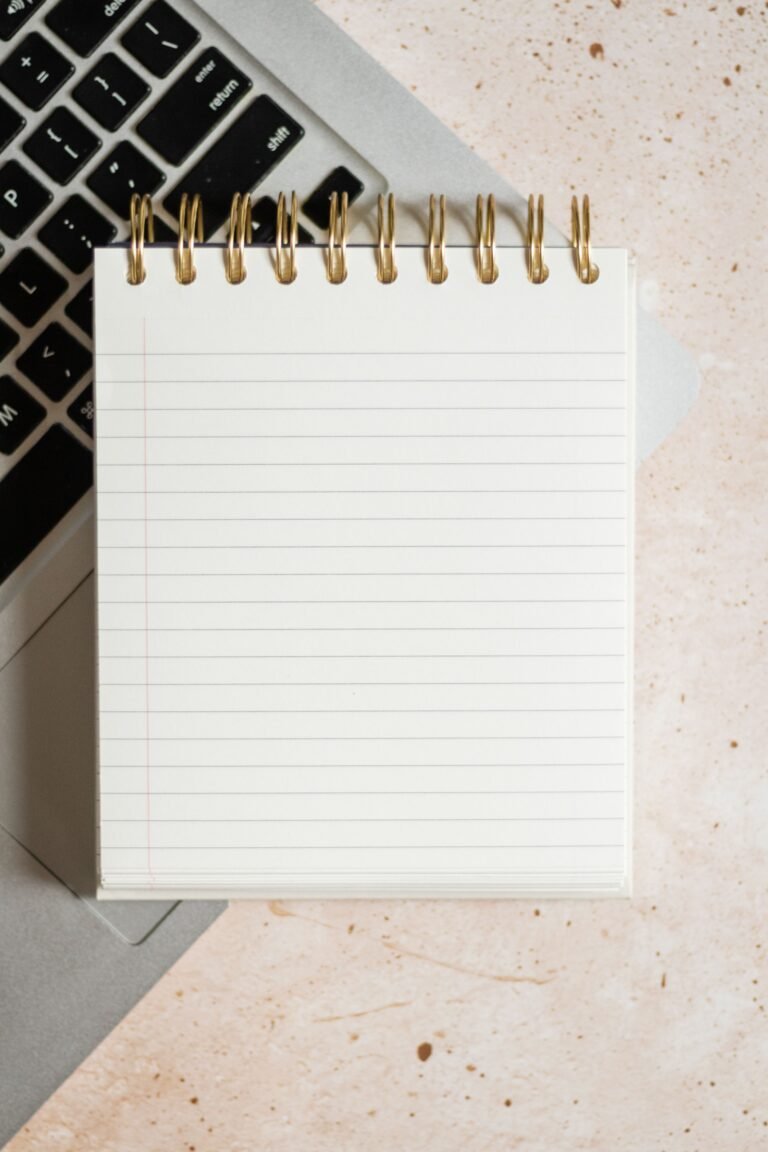 Blank spiral notepad resting on a laptop keyboard, seen from above on a speckled cream desk